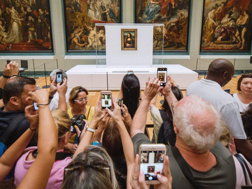 Tłum zwiedzających w muzeum przed obrazem Mona Lisa w Luwrze, fotografujący dzieło smartfonami